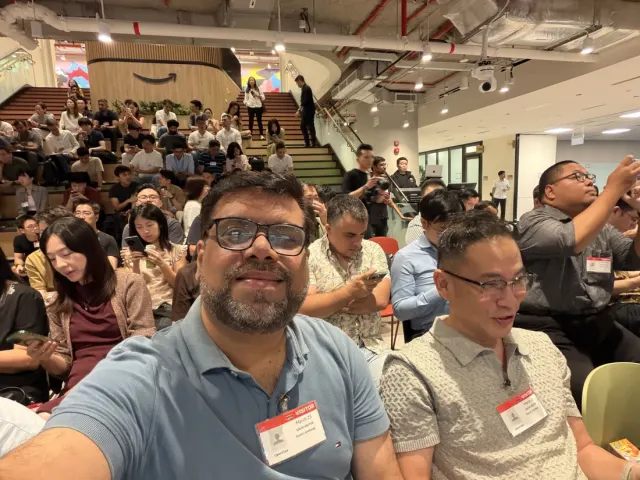 Selfie in a packed office atrium with tiered wooden seating, an Amazon logo on the wall, and an OpenClaw visitor badge