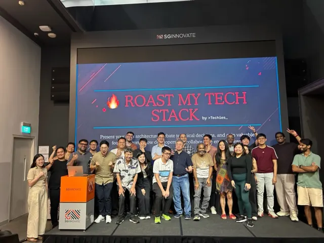 Large group posing on stage at a Roast My Tech Stack session beside an SGInnovate podium