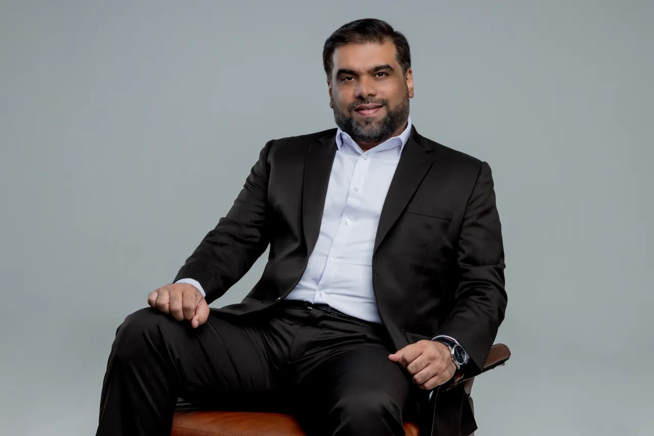 Tanveer Riaz seated in a dark suit and white shirt on a tan leather chair against a grey studio background