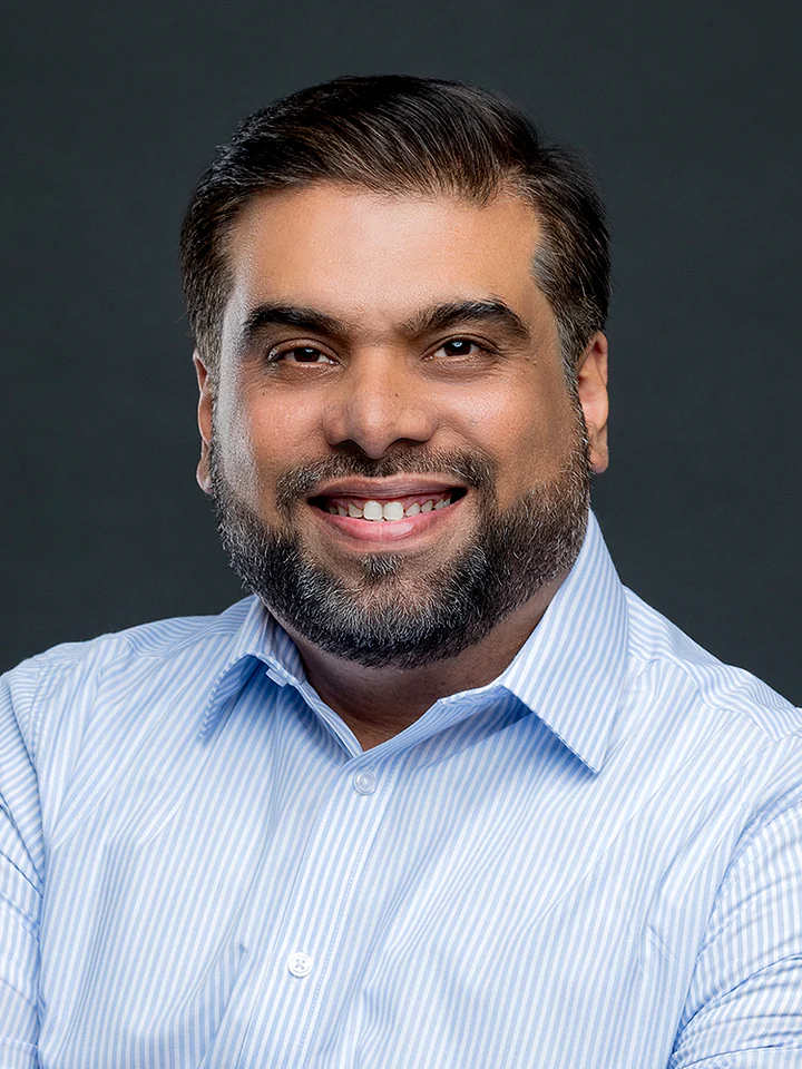 Headshot portrait of Tanveer Riaz in a blue striped shirt against a dark grey studio background