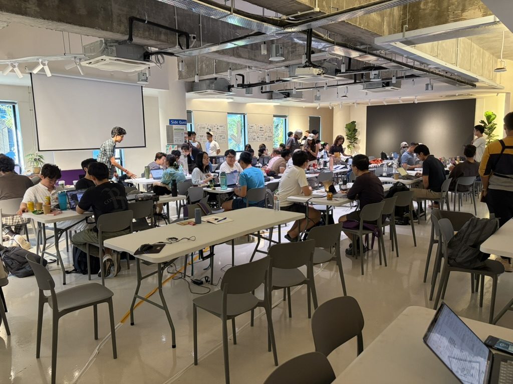Wide view of the hackathon room with side-quest board and full tables